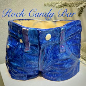 Puede incluir: Una maceta de cerámica con forma de pantalón corto vaquero azul. La maceta tiene una superficie texturizada con detalles como bolsillos, trabillas y un botón. El texto "Rock Candy Bar" está escrito en cursiva en la parte superior.