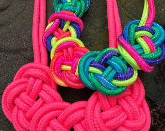 May include: A colorful rope necklace with a pink, green, yellow, and blue color scheme. The necklace is made of braided rope and has a knot design. The text "Rock Candy Bar" is visible at the top of the image.