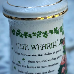 May include: White ceramic jar with a lid, adorned with gold trim. The jar features green shamrock designs and text that reads "THE WEARIN'" and a poem. A decorative item with a vintage aesthetic.