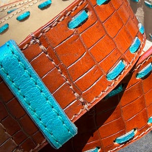 May include: Close-up of a brown leather belt with a crocodile texture. The belt features turquoise stitching and accents. The buckle strap is also turquoise. The belt is a rich brown color and appears to be handmade.