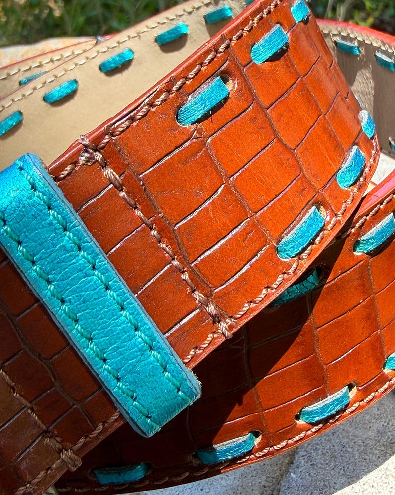 May include: Close-up of a brown leather belt with a crocodile texture. The belt features turquoise stitching and accents. The buckle strap is also turquoise. The belt is a rich brown color and appears to be handmade.
