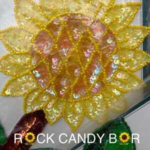 Puede incluir: Un gran aplique de girasol amarillo con un centro de cuentas y pétalos de lentejuelas. El diseño incluye una hoja marrón y una hoja verde con el texto "ROCK CANDY BOR" en blanco.