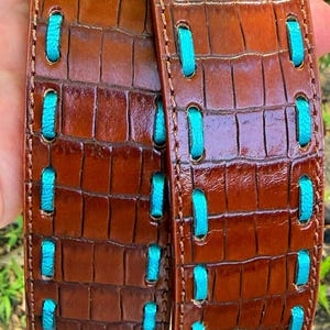 May include: Brown leather belt with a crocodile texture and turquoise stitching. The belt has a glossy finish and a repeating pattern of rectangular shapes. The turquoise stitching is visible in a series of vertical stitches.