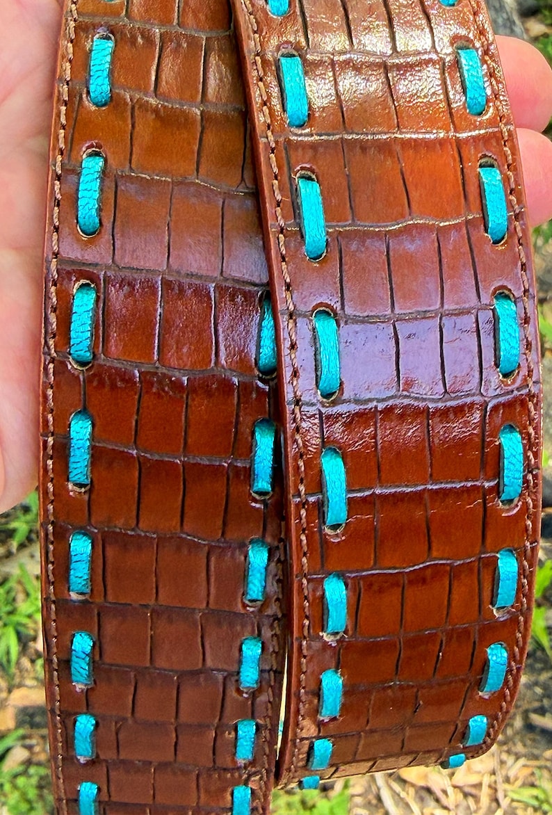 May include: Brown leather belt with a crocodile texture and turquoise stitching. The belt has a glossy finish and a repeating pattern of rectangular shapes. The turquoise stitching is visible in a series of vertical stitches.