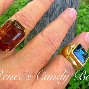 May include: Two gold rings, each with a large gemstone. One has a rectangular, amber-coloured stone, the other a square, blue stone. The rings are shown against a green backdrop, with the text "Renee's Candy Bar".