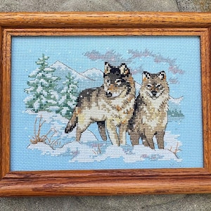 May include: Cross-stitch artwork of two wolves in a snowy landscape, framed in a wooden frame. The wolves are depicted with brown and black fur, standing in snow with a light blue background. A green pine tree and mountains are in the background.