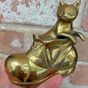 Może przedstawiać: Mosiężna figurka kota wychodzącego z buta. Kot ma widoczne detale, takie jak oczy i uszy. But ma teksturowaną powierzchnię. Całość to przedmiot dekoracyjny.