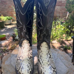 Könnte beinhalten: Ein Paar Cowboy-Stiefel mit schwarzen Lederoberteilen und einem Schlangenmuster auf dem Fuß. Die Stiefel haben dekorative Nähte und ein klassisches Western-Design. Die Stiefel werden im Freien auf einer Steinoberfläche präsentiert.
