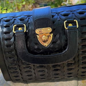 Könnte beinhalten: Schwarze Handtasche mit gewebtem Design und goldfarbener Hardware. Die Tasche hat einen oberen Griff, einen Klappenverschluss mit goldenem Verschluss und goldfarbene Akzente an den Griffbefestigungen. Die Tasche liegt auf einer Steinoberfläche.