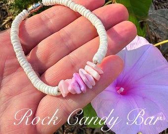 Könnte beinhalten: Ein weißes Muschel-Armband mit einem rosa und violetten Muschel-Akzent. Das Armband befindet sich auf einer Hand mit einer violetten Blume im Hintergrund.