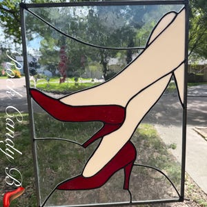 Op de afbeelding: Een rechthoekig glas-in-lood paneel met een ontwerp van benen in rode hoge hakken tegen een achtergrond van helder, getextureerd glas. Het stuk is omlijst in donker metaal en bevat de tekst Rock Candy B aan de linkerzijde.