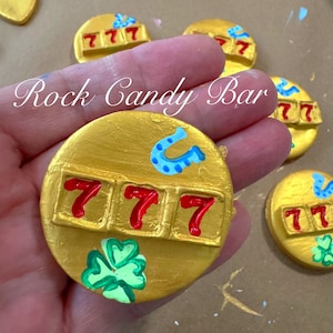 Peut inclure: Bonbon rond en pierre peint en or avec un fer à cheval, un trèfle et trois 7 rouges peints dessus. Le bonbon en pierre dit "Rock Candy Bar" en écriture cursive.