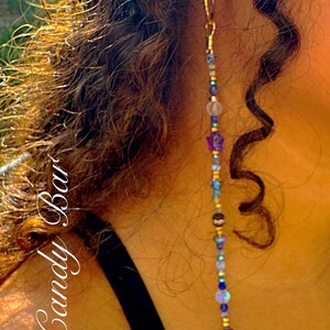 May include: A long, dangling earring with a gold hoop and a beaded strand. The strand features blue, gold, and iridescent beads, a purple star, and a crescent moon charm. The text "Rock Candy Bar" is visible.
