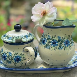 Op de afbeelding: Een blauwe en witte, met bloemen geprinte, keramische roomkan en suikerpot set met een deksel op een bijpassend dienblad. De set is versierd met blauwe bloemen en groene bladeren op een witte achtergrond.