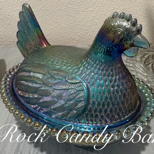 May include: Iridescent glass hen-shaped dish with a lid, featuring a detailed feather design and a beaded rim. The dish has a blue and green color with hints of purple and gold. The text "Rock Candy Bar" is at the bottom of the image.
