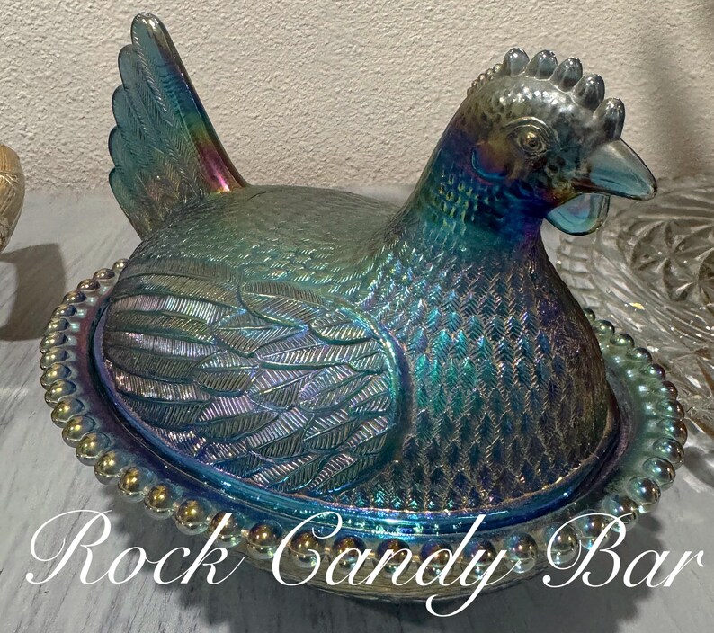 May include: Iridescent glass hen-shaped dish with a lid, featuring a detailed feather design and a beaded rim. The dish has a blue and green color with hints of purple and gold. The text "Rock Candy Bar" is at the bottom of the image.
