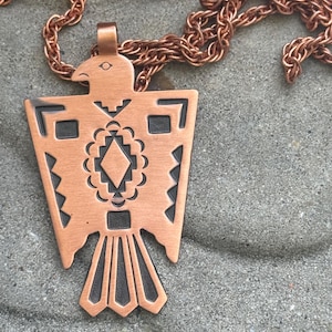 Pode incluir: Um colar com pingente de cor cobre com um design de águia estilizado. O pingente apresenta um padrão geométrico com detalhes em preto e é suspenso por uma corrente combinando. O colar é exibido em uma superfície cinza texturizada.