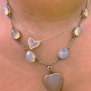 May include: A gold chain necklace with a heart-shaped pendant and multiple oval-shaped stones. The stones are a pale, milky white color. The pendant is also a pale, milky white color with a small, clear heart in the center.