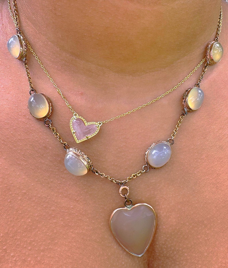May include: A gold chain necklace with a heart-shaped pendant and multiple oval-shaped stones. The stones are a pale, milky white color. The pendant is also a pale, milky white color with a small, clear heart in the center.