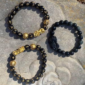 Puede incluir: Tres pulseras de cuentas negras con detalles dorados. Las pulseras presentan un dije de dragón, un dije de Buda y un dije de ojo de tigre. Las cuentas están inscritas con letras doradas.