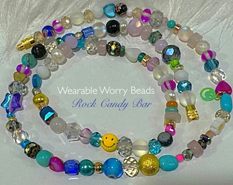 May include: A colorful beaded necklace with a variety of shapes and sizes, including stars, hearts, and smiley faces. The beads are made of glass, metal, and plastic. The necklace is called "Rock Candy Bar" and is described as "Wearable Worry Beads".