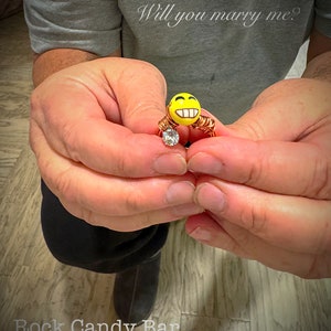 May include: A gold ring with a yellow smiley face charm and a clear diamond. The ring is held in the hands of a person. The text "Will you marry me?" is written above the ring.