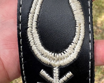 May include: Black leather strap with white embroidered designs. The designs include a horseshoe, a star, and geometric patterns.