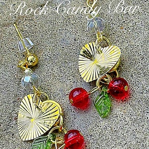 Puede incluir: Pendientes dorados con colgantes en forma de corazón con diseño de rayos de sol. Cada pendiente tiene cuentas de cereza rojas y abalorios de hojas verdes. El texto "Rock Candy Bar" es visible en la imagen.