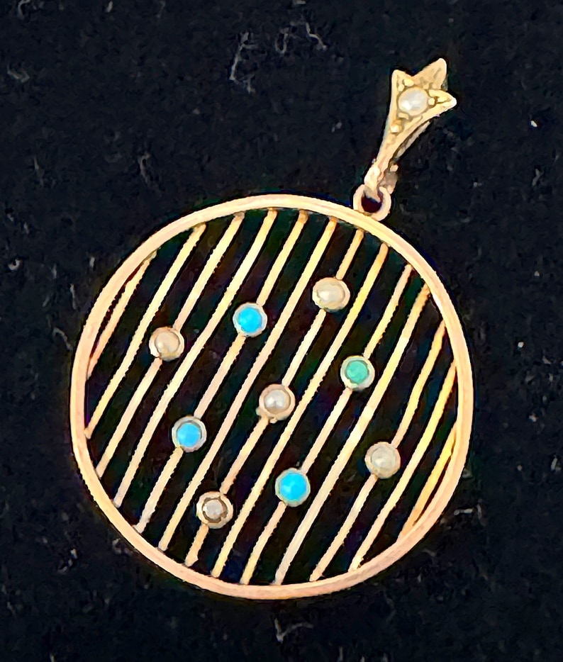 K&ouml;nnte beinhalten: Goldener Anh&auml;nger mit kreisf&ouml;rmigem Design mit parallelen Linien und abwechselnden t&uuml;rkisfarbenen und Perlenakzenten. Der Anh&auml;nger hat eine sternf&ouml;rmige Spitze mit einer Perle. Der Schmuck ist vor einem dunklen Hintergrund platziert.