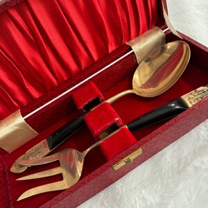 May include: A vintage cutlery set in a red velvet-lined box. The set includes a large gold-colored spoon, fork, and knife with black handles. The box has a gold clasp and a satin ribbon.