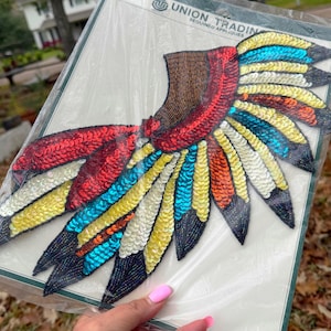 May include: A colorful sequined applique in the shape of a wing, featuring red, brown, yellow, blue, and black sequins. The applique is on a white background and is in a clear plastic package. The text "UNION TRADING SEQUINED APPLIQUES" is visible.