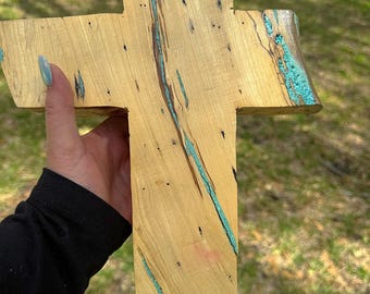 May include: A wooden cross with a turquoise blue resin inlay. The cross is made of light wood with a natural grain pattern.