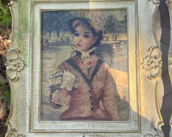 Puede incluir: Impresión enmarcada vintage que representa a una mujer en un parque, con sombrero y abrigo. El marco es ornamentado, con acabado crema y detalles dorados. La obra de arte representa a una mujer sosteniendo un objeto pequeño, con un lago y árboles al fondo.