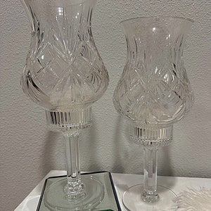 May include: Two clear glass candle holders with diamond-cut detailing. The candle holders feature a tulip-shaped top, a slender stem, and a round base. One is placed on a mirrored base with a floral design.