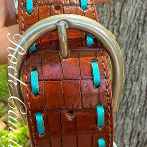 May include: Brown leather belt with a crocodile texture, accented with turquoise stitching. Features a silver buckle and turquoise trim. The belt is held up by a hand.