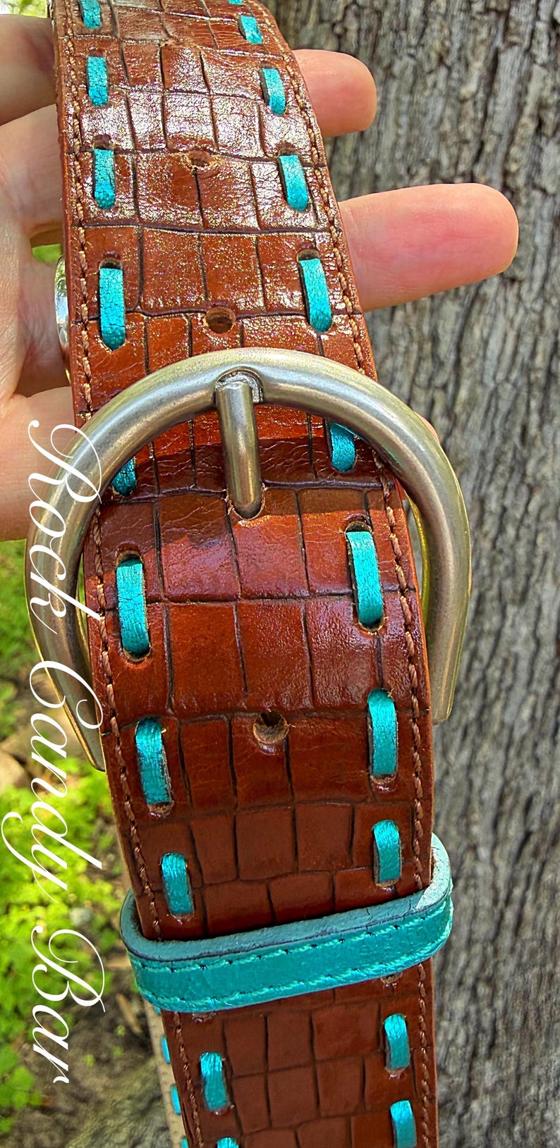 May include: Brown leather belt with a crocodile texture, accented with turquoise stitching. Features a silver buckle and turquoise trim. The belt is held up by a hand.