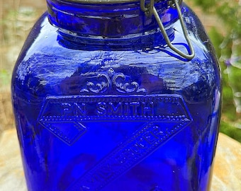 May include: A cobalt blue glass jar with a hinged lid and metal clasp. The jar has embossed text and decorative elements. The lid has a black rim and an orange rubber seal. A vintage-style container.