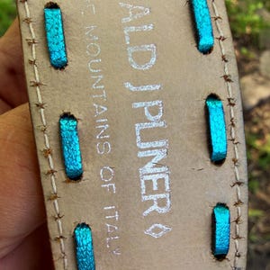 May include: Close-up of a beige leather belt with turquoise stitching and the brand name "DONALD J PLINER" in silver. The text "MOUNTAINS OF ITALY" is also visible. The belt has a stitched border.