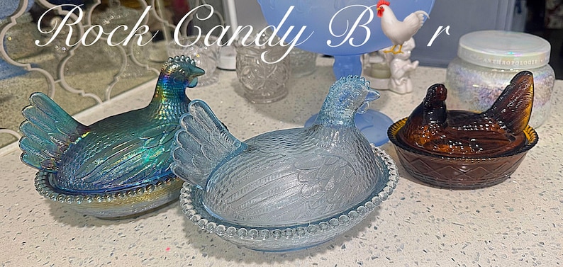 May include: Three glass hen-shaped dishes in iridescent blue-green, light blue, and brown. The dishes are sitting on a white surface. The text "Rock Candy Bar" is at the top of the image.