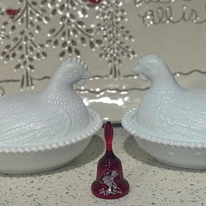 May include: Two white ceramic hen-shaped covered dishes with textured details, sitting on a white surface. A small, red bell with a floral design is between them. A Santa Claus figurine is visible on the left.