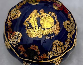May include: A blue and gold porcelain trinket box with a lid featuring a couple in a garden scene. The box is decorated with gold floral designs and the word "Limoges" is inscribed on the lid.