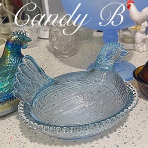 May include: Three glass hen-shaped dishes in iridescent blue, clear, and brown. The hens sit atop round, beaded bases. The text "Rock Candy Bar" is at the top of the image. A blue pedestal dish and a jar with a white lid are in the background.