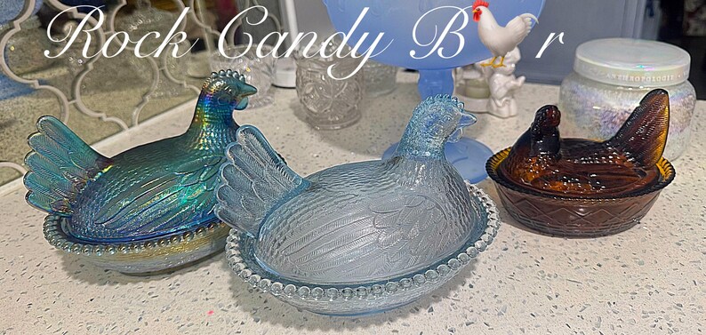 May include: Three glass hen-shaped dishes in iridescent blue, clear, and brown. The hens sit atop round, beaded bases. The text "Rock Candy Bar" is at the top of the image. A blue pedestal dish and a jar with a white lid are in the background.