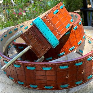May include: A brown leather belt with a crocodile texture and turquoise stitching. The belt has a silver buckle and a turquoise leather loop. The belt is approximately 1.5 inches wide.
