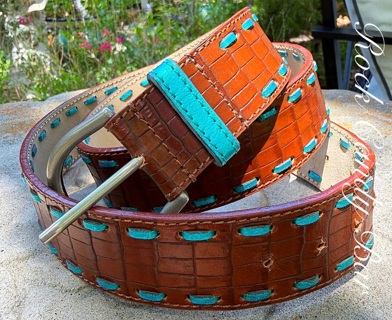 May include: A brown leather belt with a crocodile texture and turquoise stitching. The belt has a silver buckle and a turquoise leather loop. The belt is approximately 1.5 inches wide.