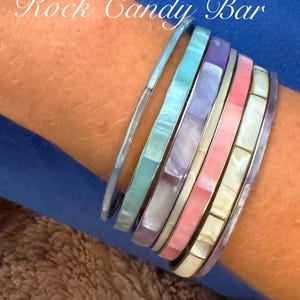 Op de afbeelding: Een set van zes kleurrijke armbanden met een parelmoerontwerp. De armbanden zijn gestapeld op de pols van een persoon en zijn gemaakt van verschillende materialen, waaronder metaal en plastic. De armbanden zijn in tinten blauw, paars, roze en wit.