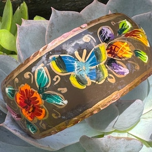 Op de afbeelding: Een bruine metalen armband met handgeschilderde vlinders en bloemen. De vlinders zijn blauw, geel en groen, en de bloemen zijn rood, oranje en geel.