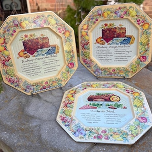 May include: Three octagonal decorative plates with a cream background and a colourful fruit and floral border. Each plate features a different dessert recipe: Blueberry-Orange Nut Bread and Buche de Noel. The plates are displayed on a stone surface.