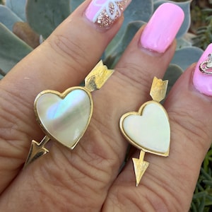 May include: Two gold-toned rings, each featuring a heart-shaped, iridescent mother-of-pearl center. The hearts are pierced by a gold arrow. The rings are displayed on a hand with pink nail polish.