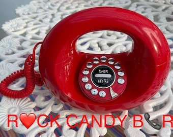 May include: A bright red circular landline telephone with a glossy finish, featuring a central circular keypad with silver buttons, a red coiled cord, and the text "ROCK CANDY BAR" printed in red at the bottom of the frame with a heart and phone icon.